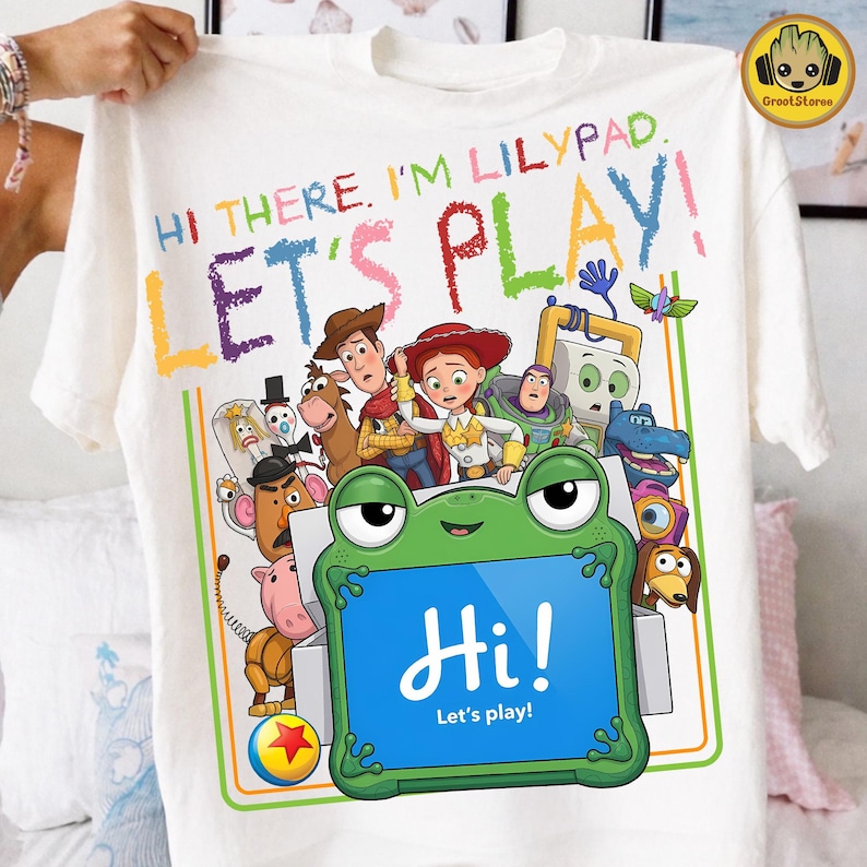 May include: A white t-shirt featuring a colorful graphic of Toy Story characters and a green frog-shaped tablet. The text reads: HI THERE, I'M LILYPAD. LET'S PLAY! and Hi! Let's play! The design includes Woody, Jessie, Buzz Lightyear, and others.