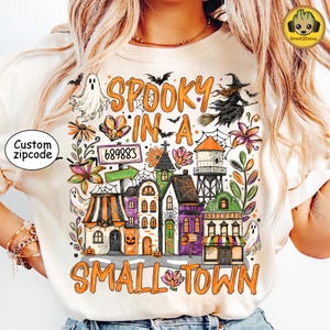 May include: A cream-colored t-shirt featuring a colorful Halloween-themed graphic with the text "SPOOKY IN A SMALL TOWN" and "689883". The design includes a ghost, a witch on a broom, a water tower, various buildings, spiderwebs, and flowers.
