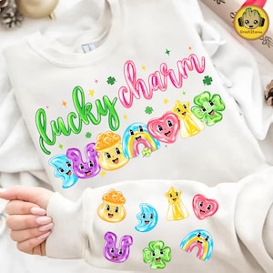 Puede incluir: Sudadera blanca con las palabras "lucky charm" en letras verdes y rosas. El diseño presenta amuletos de dibujos animados coloridos, incluyendo una herradura, un arco iris, un corazón, una estrella y un trébol. Hay amuletos adicionales dispersos debajo.