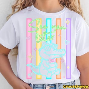 May include: White t-shirt featuring a neon alligator wearing sunglasses and a bow tie, with the text "See you later." Vertical stripes in pink, blue, orange, yellow, and purple are behind the alligator.