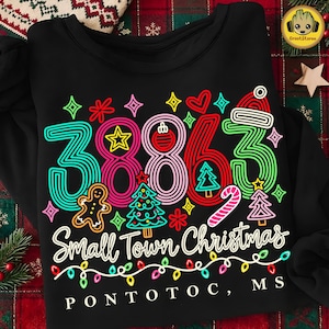 May include: A black crewneck sweatshirt featuring the number 38863 in a colourful, retro-style striped font. The design includes Christmas icons like a gingerbread man, trees, a candy cane, and a Santa hat, with the text Small Town Christmas and PONTOTOC, MS.