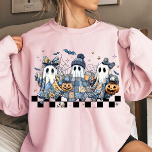 K&ouml;nnte beinhalten: Hellrosa Sweatshirt mit einem Halloween-Motiv. Das Design zeigt drei Geisterfiguren, die K&uuml;rbiseimer halten und H&uuml;te und Schleifen tragen. Flederm&auml;use, Sterne und Spinnweben erg&auml;nzen die Illustration. Am unteren Rand befindet sich ein schwarz-wei&szlig; karierter Rand.