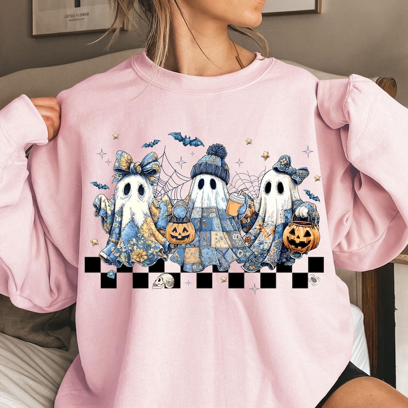 K&ouml;nnte beinhalten: Hellrosa Sweatshirt mit einem Halloween-Motiv. Das Design zeigt drei Geisterfiguren, die K&uuml;rbiseimer halten und H&uuml;te und Schleifen tragen. Flederm&auml;use, Sterne und Spinnweben erg&auml;nzen die Illustration. Am unteren Rand befindet sich ein schwarz-wei&szlig; karierter Rand.