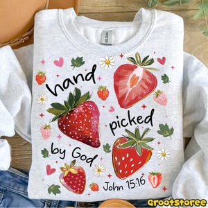 May include: A light grey sweatshirt featuring a strawberry design and the text "hand picked by God John 15:16". The design includes red strawberries, daisies, and hearts. The sweatshirt is a Gildan brand.