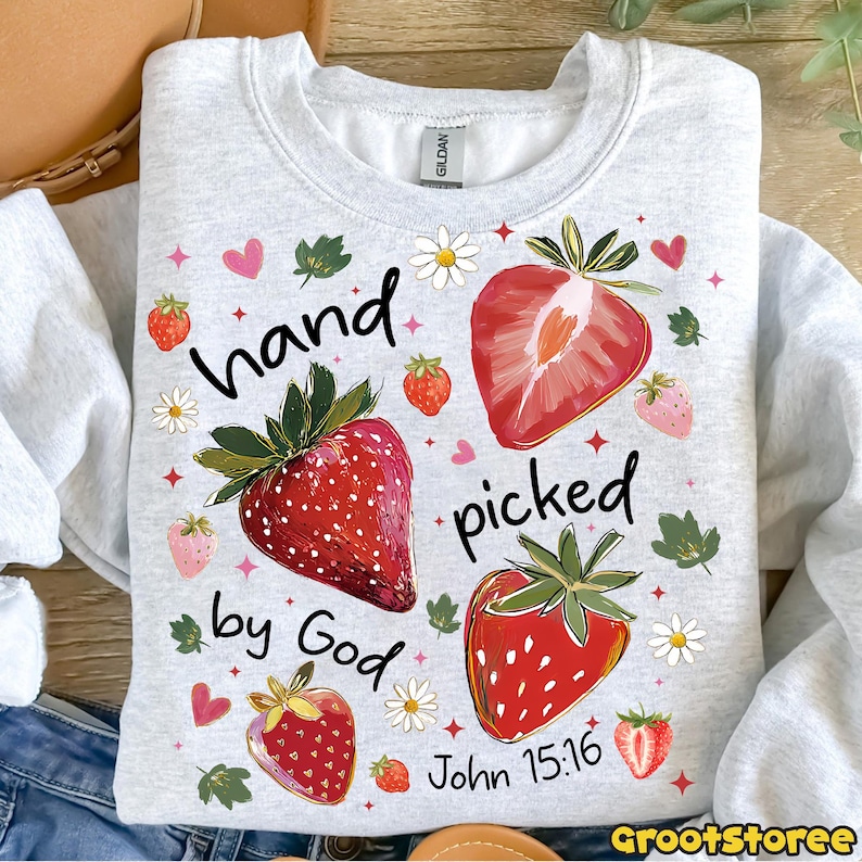 May include: A light grey sweatshirt featuring a strawberry design and the text "hand picked by God John 15:16". The design includes red strawberries, daisies, and hearts. The sweatshirt is a Gildan brand.