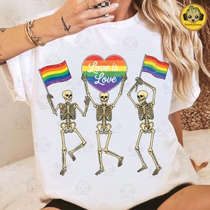 May include: White t-shirt featuring three skeleton figures holding rainbow flags and a heart-shaped sign that reads "Love is Love." The skeletons are in a dancing pose, with the flags and heart in the air. The design is a colorful celebration of love and inclusivity.