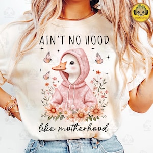 May include: A white t-shirt with a pink duck wearing a pink hoodie and surrounded by flowers. The text "Ain't no hood like motherhood" is printed on the shirt.
