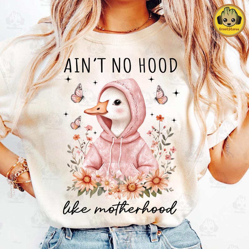 May include: A white t-shirt with a pink duck wearing a pink hoodie and surrounded by flowers. The text "Ain't no hood like motherhood" is printed on the shirt.