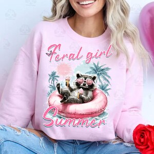 May include: Pink sweatshirt with a graphic of a raccoon wearing sunglasses and holding an ice cream cone. The raccoon is sitting in a pink inflatable ring. The text "Feral girl summer" is printed on the sweatshirt.