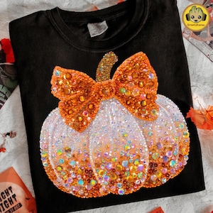 May include: Black sweatshirt featuring a sparkling pumpkin design. The pumpkin is white with an ombre effect, transitioning to orange at the bottom. An orange bow with gold glitter accents sits atop the pumpkin. The design is embellished with colorful rhinestones.