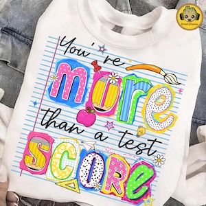 May include: White sweatshirt with a colorful graphic that reads "You're more than a test score." The design includes an apple, pencil, and other school-themed elements. The text is in various bright colors.