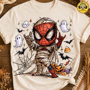 May include: A cream-colored t-shirt featuring a graphic of a character dressed as a mummy with a red and black mask, making a rock-and-roll hand gesture. The design includes white ghosts, black bats, orange pumpkins, and candy corn illustrations.