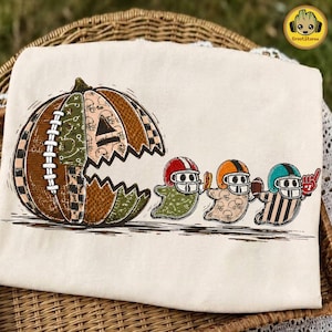 May include: A cream-colored t-shirt featuring a graphic of a large pumpkin designed like a video game character, with a mouth open toward three ghost figures wearing football helmets. The ghosts hold a hot dog, a football, and a foam finger.