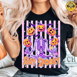 Peut inclure: Un t-shirt noir avec un motif de squelette violet assis en position du lotus, entouré de citrouilles orange, de petites chauves-souris violettes et d'étoiles sur un fond rayé rose et noir avec le texte Stay Spooky écrit en orange.