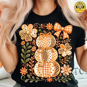 May include: A black t-shirt featuring a graphic print of three stacked pumpkins decorated with orange and white gingham, leopard print, and floral patterns. The design includes orange flowers, green leafy vines, and an orange gingham bow.