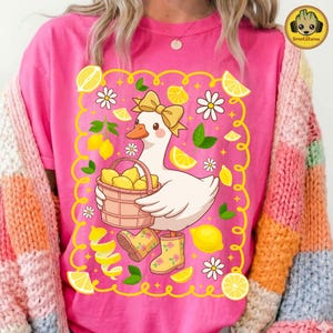 May include: Pink t-shirt featuring a cartoon goose wearing a bow, holding a basket of lemons, and wearing yellow rain boots. The design includes lemons, daisies, and lemon peel accents, all within a yellow border.