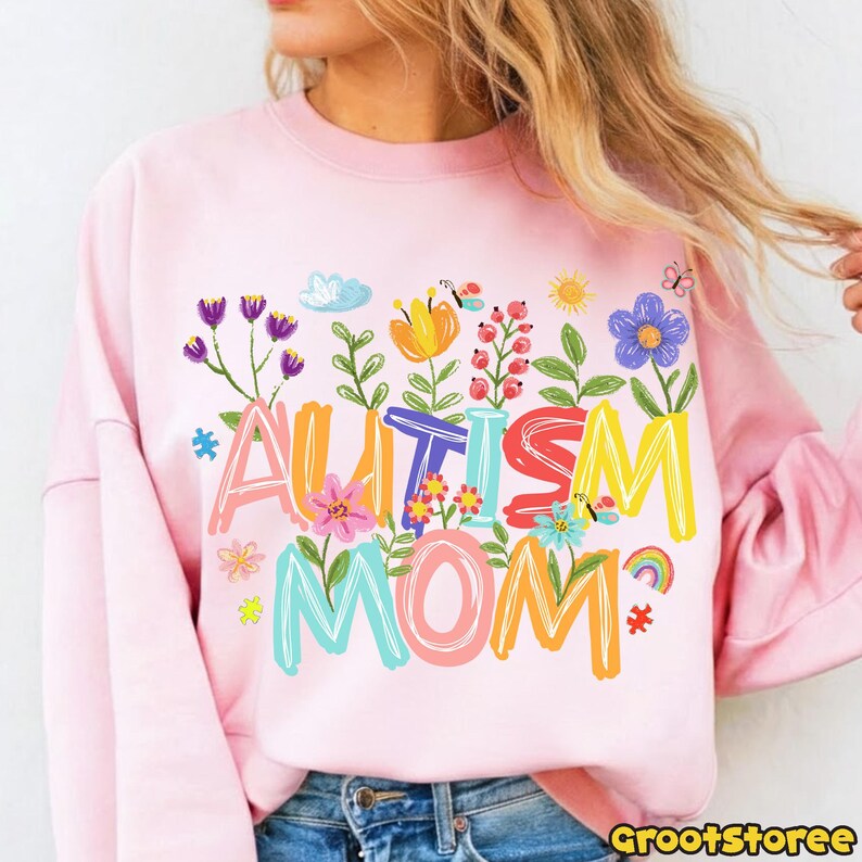 Puede incluir: Sudadera rosa claro con las palabras "AUTISM MOM" en letras coloridas y florales. El dise&ntilde;o incluye flores, un arco&iacute;ris y piezas de rompecabezas, creando un mensaje alegre y de apoyo.