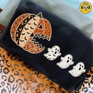 May include: A black sweatshirt featuring a large pumpkin graphic divided into segments with patterns of jack-o'-lanterns, candy corn, bats, and skulls. Three white ghost figures with black eyes and mouths are positioned to the right of the pumpkin.