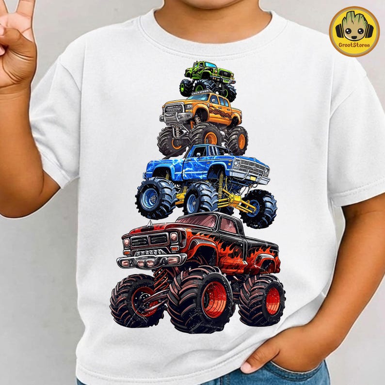 May include: White t-shirt featuring a vibrant graphic of five monster trucks stacked on top of each other. The trucks are in various colors, including red, orange, blue, and green, with large tires and detailed designs. The shirt has a crew neck.