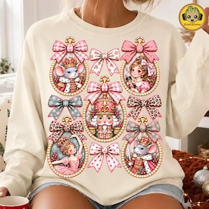Può includere: Una felpa girocollo color crema con una stampa grafica di cinque ornamenti ovali e sei fiocchi decorativi. Gli ornamenti contengono illustrazioni di un re topo, una ragazza, uno schiaccianoci, una ballerina e un topo soldato in rosa e oro.