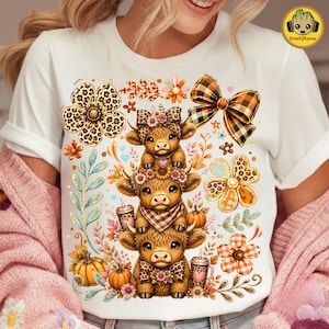 Può includere: Una t-shirt bianca con una stampa grafica di tre mucche Highland impilate. Il design include un fiore con stampa leopardata, un fiocco a quadri, zucche arancioni e vari elementi floreali autunnali attorno alle mucche impilate.