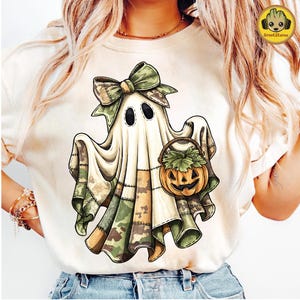 May include: Cream-colored t-shirt featuring a cartoon ghost with a camouflage bow and patchwork design. The ghost holds a jack-o'-lantern bucket. The design incorporates shades of green, brown, and orange, perfect for Halloween.