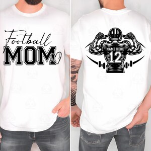 May include: White t-shirt with the words "Football MOM" in black script and block letters, with a football-shaped heart graphic. The back of the shirt features a football player graphic with the text "NAME HERE" and the number "12".