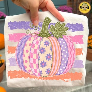 May include: A white t-shirt featuring a large pumpkin graphic with alternating segments of purple polka dots, pink and white checkers, purple flowers, and orange stripes. The pumpkin has a green stem and leaf, set against horizontal brushstroke stripes.