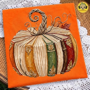 Peut inclure: Un t-shirt orange avec un motif de citrouille form&eacute;e par des livres empil&eacute;s. La citrouille poss&egrave;de une tige tress&eacute;e, des vrilles recourb&eacute;es et des feuilles d'automne. Les dos des livres sont color&eacute;s en jaune, vert, marron et rouge avec des d&eacute;tails dor&eacute;s.