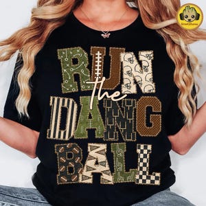 May include: A black t-shirt featuring the text RUN THE DANG BALL in large, multi-patterned block letters. The letters incorporate various football-themed designs including play diagrams, helmets, stripes, footballs, pennants, and foam fingers.