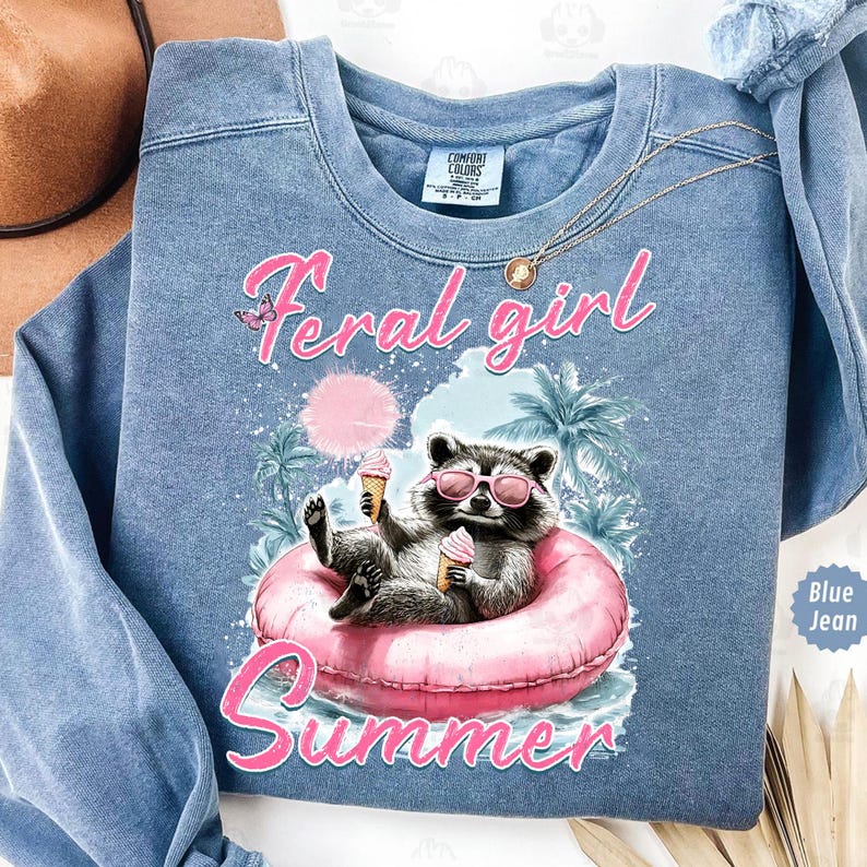 May include: A blue jean sweatshirt with the text "Feral girl summer" and a cartoon raccoon wearing sunglasses and holding an ice cream cone in a pink inflatable ring.