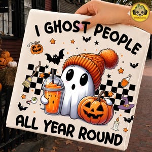 Op de afbeelding: Een witte draagtas met een cartoongeest die een oranje gebreide muts draagt, een oranje drankje vasthoudt en naast een pompoen zit. De tekst luidt I GHOST PEOPLE ALL YEAR ROUND, omringd door vleermuizen, sterren en een dambordpatroon.