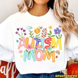 Puede incluir: Sudadera blanca con las palabras "AUTISM MOM" en letras coloridas, decorada con flores, mariposas y un arco&iacute;ris. El dise&ntilde;o incluye piezas de rompecabezas y un sol. Un dise&ntilde;o alegre y solidario.