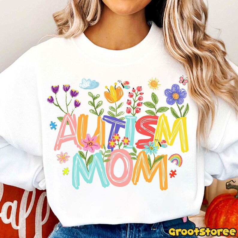 Puede incluir: Sudadera blanca con las palabras "AUTISM MOM" en letras coloridas, decorada con flores, mariposas y un arco&iacute;ris. El dise&ntilde;o incluye piezas de rompecabezas y un sol. Un dise&ntilde;o alegre y solidario.