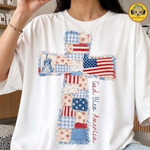 May include: White t-shirt featuring a cross design made of patchwork fabric. The cross incorporates elements like the American flag, Statue of Liberty, and floral patterns. The text "God Bless America" is written vertically.