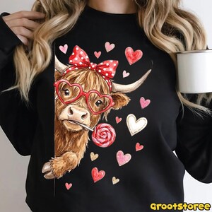May include: Black sweatshirt featuring a brown Highland cow wearing heart-shaped glasses and a red polka dot bow. The cow is holding a lollipop and surrounded by pink and white hearts. The text "grootstoree" is at the bottom.