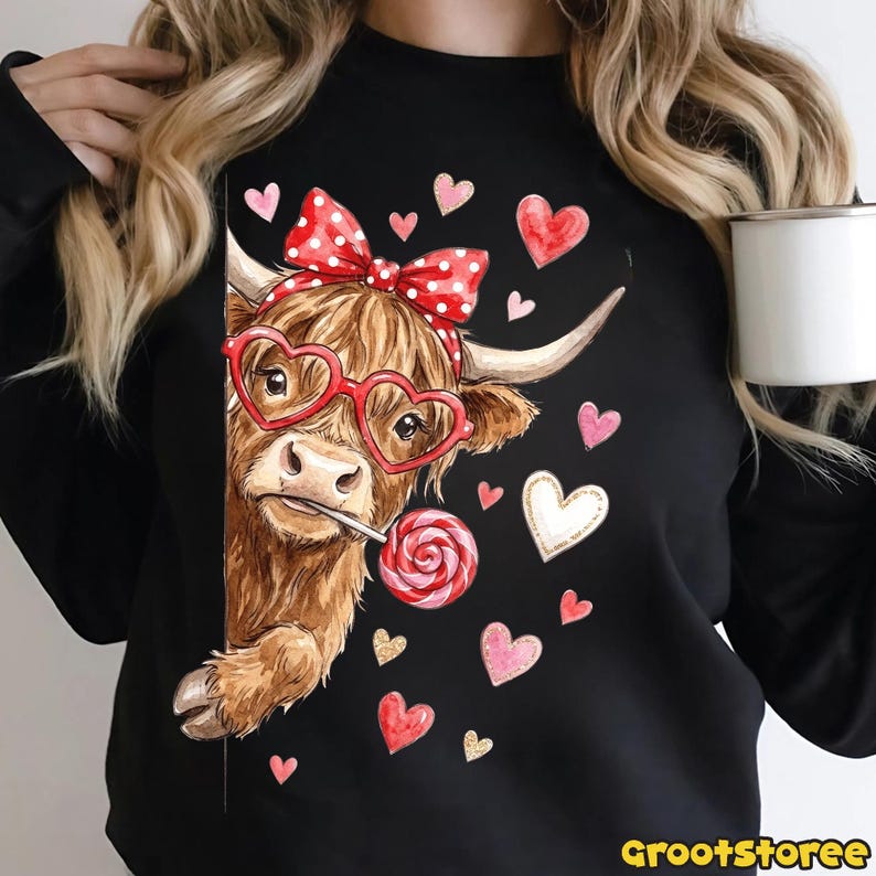 May include: Black sweatshirt featuring a brown Highland cow wearing heart-shaped glasses and a red polka dot bow. The cow is holding a lollipop and surrounded by pink and white hearts. The text "grootstoree" is at the bottom.