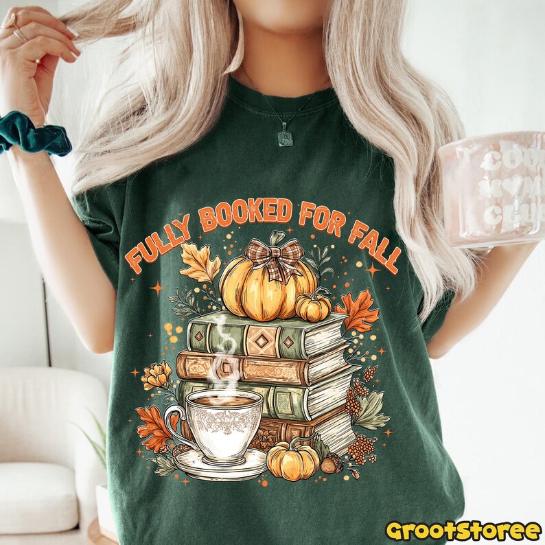 May include: Dark green t-shirt with the text "FULLY BOOKED FOR FALL" above a graphic of pumpkins, books, and a cup of coffee. The design features autumn leaves and a bow. The shirt has a crew neck and short sleeves.