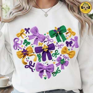 May include: White sweatshirt featuring a heart-shaped design of crocheted bows in purple, yellow, and green. The bows are intricately detailed, with a variety of sizes and styles. The design includes small heart and bow accents.