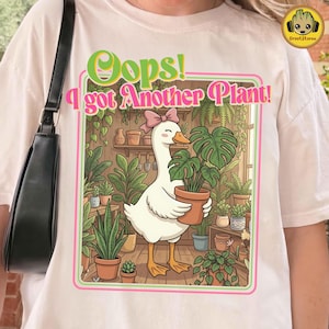 May include: White t-shirt with a graphic featuring a cartoon goose holding a potted plant. The design includes the text "Oops! I got Another Plant!" in green and pink, surrounded by a pink and green border. The goose is in a greenhouse setting.