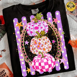 May include: A black t-shirt featuring a graphic of three stacked pumpkins inside a gold oval frame. The top pumpkin is light purple with pink spots, the middle has a leopard print, and the bottom has a pink and white checkerboard pattern.