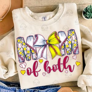 May include: A beige sweatshirt with the text "Mom of both" in maroon letters. The letters are decorated with a yellow and white softball bow and softball designs.