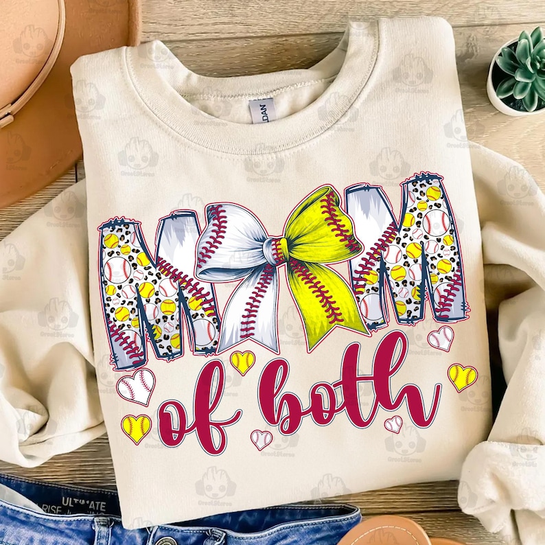 May include: A beige sweatshirt with the text "Mom of both" in maroon letters. The letters are decorated with a yellow and white softball bow and softball designs.