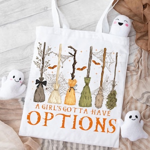 May include: White tote bag featuring a whimsical design of various witch brooms, spiderwebs, and bats. The text "A Girl's Gotta Have Options" is printed in orange. The bag has white handles and is decorated with plush ghost figures.