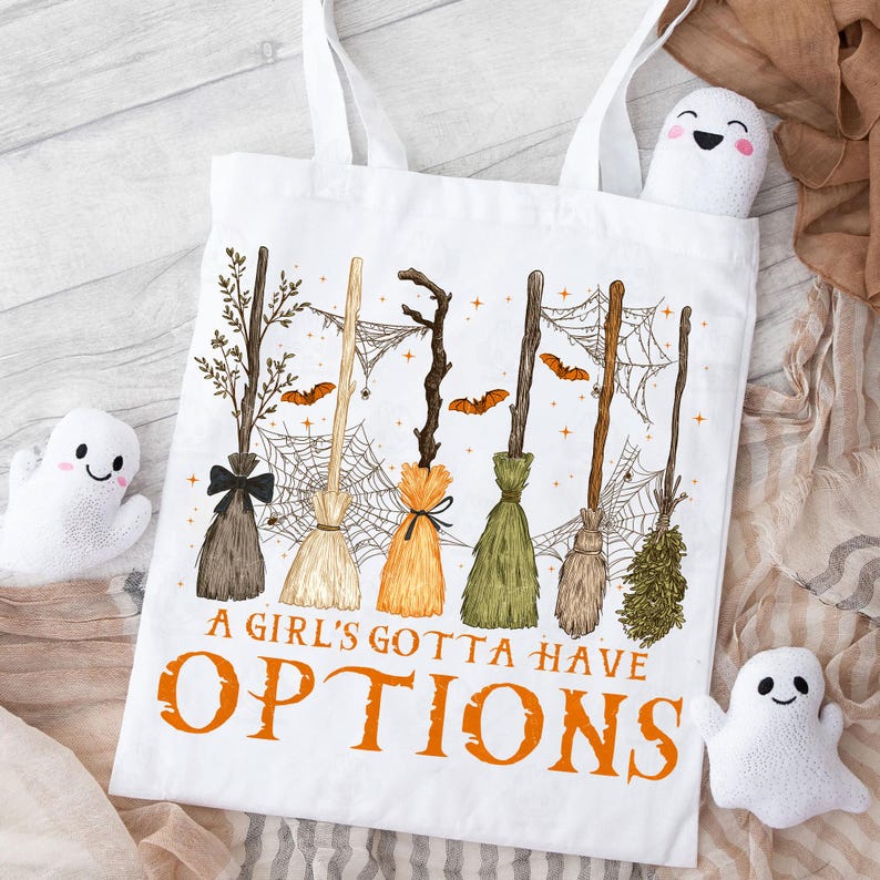 May include: White tote bag featuring a whimsical design of various witch brooms, spiderwebs, and bats. The text "A Girl's Gotta Have Options" is printed in orange. The bag has white handles and is decorated with plush ghost figures.