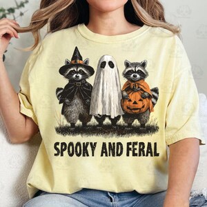 May include: Yellow t-shirt with a Halloween-themed graphic. The design features three raccoons dressed as a witch, a ghost, and holding a pumpkin. The text "Spooky and Feral" is printed below the characters.