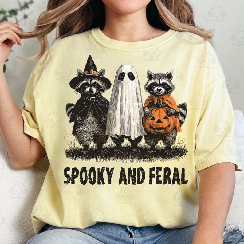 May include: Yellow t-shirt with a Halloween-themed graphic. The design features three raccoons dressed as a witch, a ghost, and holding a pumpkin. The text "Spooky and Feral" is printed below the characters.