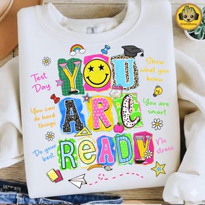 Könnte beinhalten: Weißes Sweatshirt mit einem farbenfrohen Aufdruck, der "YOU ARE READY" mit verschiedenen schulbezogenen Illustrationen zeigt. Das Design beinhaltet ein Smiley, eine Doktorhut und den Text "Test Day."