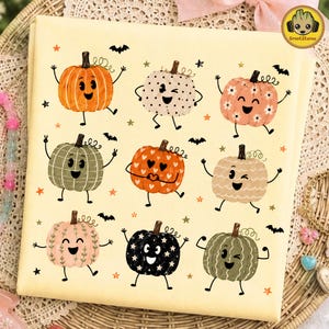 May include: A light yellow fabric square featuring a repeating pattern of nine cartoon pumpkins with faces, arms, and legs. The pumpkins are decorated with various patterns including polka dots, flowers, hearts, and stars, interspersed with small black bats and orange stars.
