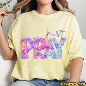 May include: A light yellow t-shirt with the words "just PRAY" in a floral design. The word "just" is in cursive, and the word "PRAY" is in large, colorful letters with flower accents. The shirt has a crew neck.